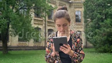 年轻漂亮的女人穿着五颜六色的夹克，在平板电脑上浏览，<strong>坐在</strong>大学校园附近的<strong>公园里</strong>，迷人而迷人